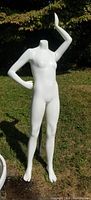 Matte white junior female mannequin with arms posed and no head, standing on a rusted metal stand outdoors on grass.