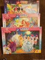 Three boxes of Disney double-face puzzles arranged stacked, showing characters Mickey Mouse, Minnie, Ariel and Cinderella with pumpkin carriage.