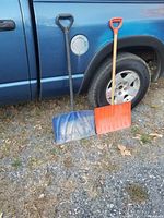 Two used snow shovels placed standing against a blue vehicle wheel, one black-handled with a blue scoop, the other orange-handled with an orange scoop.