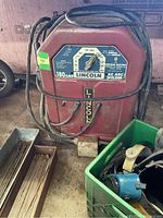 Front view of the red Lincoln Electric AC-180-S arc welder with cords wrapped around it, trays with welding rods to the side, and a green crate with helmet and glasses.