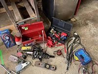 Overview of lot showing black scissor car jack, red toolbox, ratchet straps, battery charger, couplers, tire wrenches, and funnel on garage floor.
