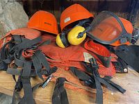 Two orange Stihl helmets with mesh face shields, yellow earmuffs and orange protective pant leg covers with straps, all showing visible wear.