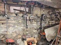 Three STIHL string trimmers hanging on stone wall with visible orange engine housings and silver shafts, box of miscellaneous parts at floor.