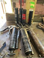 Three used metal manual grease guns laid out on surface, showing wear, including one with attached flexible hose.
