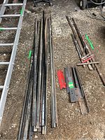Overall view of metal rods, pry bars, rebar, and fence posts laid on ground by a ladder showing size and condition with wear and rust