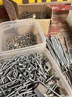 Top-down view of assorted nails and spikes in two plastic containers and one cardboard box, showing variety of nails including spiral and spike types, some with rust.