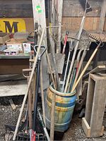 Angled view of multiple garden tools including shovels and rakes stored inside a painted ceramic barrel with wooden handles showing wear from use.