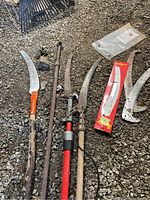 Overview photo of various pole saws and gardening tools displayed on gravel ground including saws, new blade in packaging, garden auger and rake.