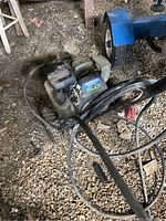 Overall view of the pressure washer on a gravel surface with hoses visible. Shows dirt and wear on the engine and frame.