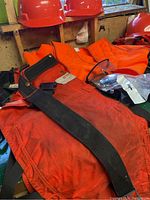 Two orange hard hats, an orange quilted safety vest, orange protective chaps with black strap and wear marks, and clear safety glasses on a surface near a window.