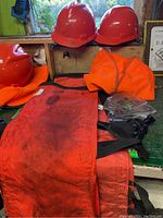 Three red hard hats, orange safety vest, orange chaps, and clear safety glasses arranged on a bench with visible dirt on fabric items.