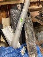 Various rolls of material including landscape cloth, vinyl tarp, plastic snow fencing, and paper, shown stored in a shed or workshop.