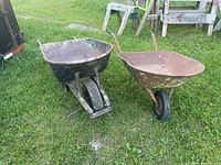 Two wheelbarrows on grass. The larger has wooden frame and black metal bucket; smaller is fully metal and rusted.