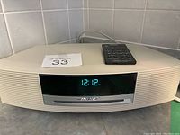 Front view of beige Bose Wave music system console showing digital clock display set to 12:12 and built-in CD slot with remote on top