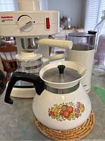 Photo of the Multichef espresso/cappuccino maker with its dial, red power button, steam wand, and clear glass pot alongside the floral Corning Ware tea kettle with black handle and lid knob.