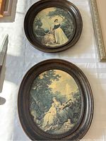 Two oval framed vintage lithographs shown face up on surface, detailed pastoral scenes in wood frames.