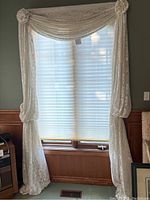 Pair of white lace window drapery panels fitted over a window with white blinds. Panels have a swag style top with rosette fabric accents and are tied at mid-length on the sides.