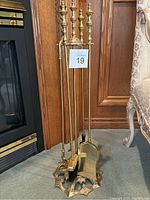Full view of vintage brass fireplace tool set leaning against wooden paneling near a fireplace.