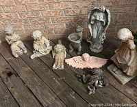 Overview of all garden figurines and decor items arranged on wood deck near brick wall, showing variety of materials and weathered condition.