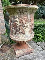 Front view of the terracotta garden urn with detailed cherub bas-relief and ornamental bands. Shows weathering and moss growth at top rim.
