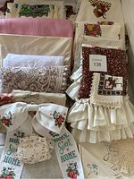 Full assortment of folded tablecloths stacked showing different colors and lace patterns, plus embroidered bow and doily.
