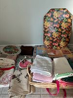 Wide view showing assorted kitchen linens including placemats, napkins, and tea towels arranged on the floor and in a basket with a floral decorative tray standing behind.