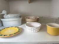 Various kitchenware items on white shelf including white ceramic duck covered dish, Sovereign Potters earthenware bowls, yellow pottery bowl, and glass corncob dishes.