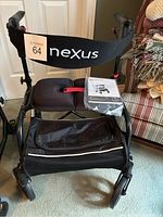 Full view of black Nexus walker with padded seat, backrest strap labeled Nexus, storage bag, and user manual on seat.