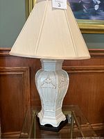 Pair of white ceramic table lamps with beige fabric shades and wood bases on glass table in front of wood paneled wall