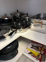 Various pots and pans on a corner shelf including saucepans with lids, aluminum roasting pans, round egg poacher pan, and perforated steaming insert.