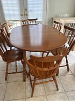 Photo showing the wooden dining table surrounded by six wooden chairs, with the leaf extension inserted making the table oval-shaped.