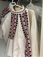 White long-sleeve Ukrainian embroidered top with red and black geometric embroidery on sleeves, chest, and neckline hung on a hanger with other garments behind it