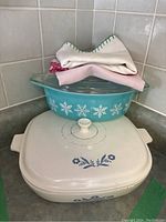 Two Pyrex casserole dishes stacked: turquoise with snowflake design (top) and white with blue floral design (bottom). Cloths are stacked over the turquoise dish lid.