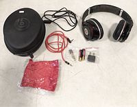 Photo showing Beats headphones with ear pads and headband, hard carrying case with Beats logo, coiled red audio cable, batteries, and adapters.