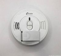 Top view photo of a white round Kidde smoke alarm with hush/test button and front loading battery compartment.