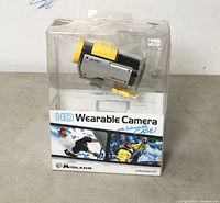 Front view of Midland HD Wearable Camera sealed in plastic retail box. Shows yellow lens cover and black body inside a clear protective case.