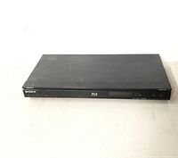 Front view of black Sony Blu-ray DVD player on white surface against white background. Player has front display and power button.