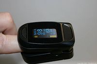 Close-up photo showing the pulse oximeter on a finger with display showing 98% SpO2, pulse rate 63 bpm, and a pulse bar graph.