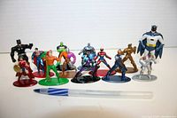 Front angle photograph of 11 superhero diecast figurines on colorful bases alongside a pen for scale.