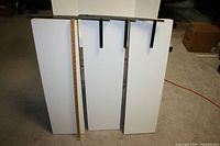 Three white floating shelves photographed upright with three metal mounting brackets visible on backs. A wooden ruler placed alongside shows shelving height approx. 36 inches.