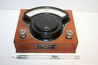 Front-right angled view showing the curved analog dial on black casing mounted on wooden base with metal terminals and branding plate.