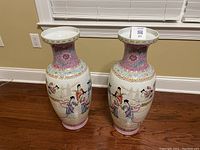 Two tall Chinese porcelain vases with pink and multicolored hand-painted traditional scenes, placed on hardwood floor under window.