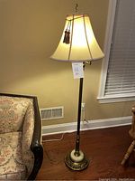 Photo of antique floor lamp with marble base, cream fabric lampshade with tassels, turned on, standing near a floral upholstered sofa and wood floor.