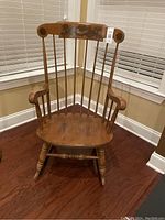 Photo of an antique wooden rocking chair standing on a wooden floor in a corner by windows with blinds. Shows front view with visible weathering on the wood finish.