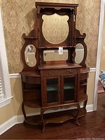 Front view of antique wooden whatnot hutch showing the large central mirror, two smaller side mirrors, glass-front cabinet doors, and curved legs.