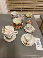 Photo showing seven assorted porcelain tea cups, saucers, and tea bag holders with floral and decorative patterns on a gray table.