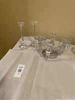 Photo of a pair of tall glass candle sticks and an antique-style crystal multi-holder with three candle holders and two side vases, all resting on a white surface.