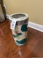 Chinese porcelain umbrella stand with floral lotus motif, glass top, positioned against wall on wooden floor, showing cylindrical shape and detailed painted decoration.