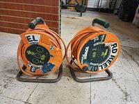 Two orange extension cord reels on metal stands, positioned side by side on floor, front view showing cords wrapped and exposed plugs