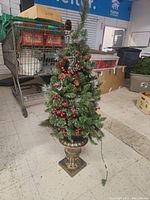 Photo of small artificial Christmas tree in decorative gold urn base with pine cones and red berries in a room with boxes and shopping cart
